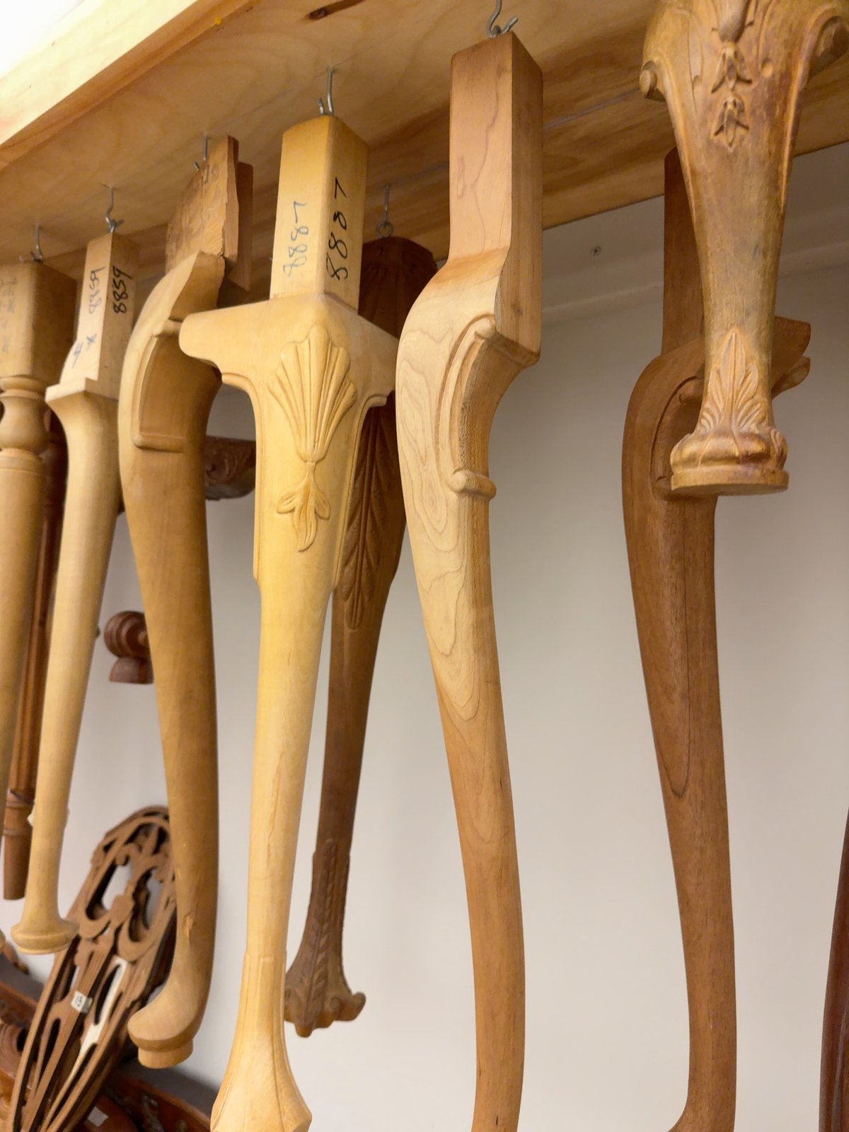 A wood shelf hung with carved chair-leg samples in unfinished hardwood: cabriole legs with various carved feet (ball-and-claw, scroll, paw), a turned spindle leg, and a square-tapered leg with reeded shaft. Each leg-top is a small square block caught on a metal hook, with a hand-written SKU tag.
