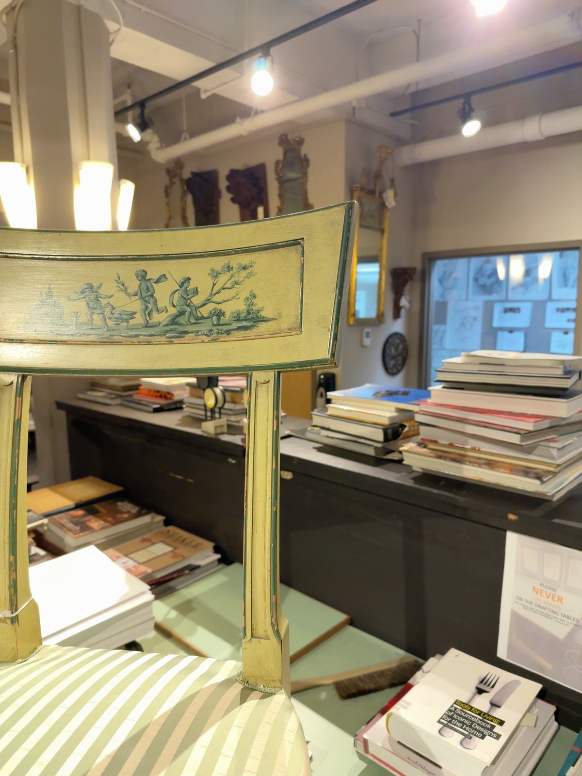 A small painted side chair viewed from the front-left. Cream-painted wood with darker green trim along every edge. The back is a single sweeping panel painted with a green-on-cream pastoral scene of figures with hounds among classical trees. The seat is upholstered in narrow cream-and-green stripes. The reeded square front leg shows wear along its edges where the paint has rubbed through to wood.