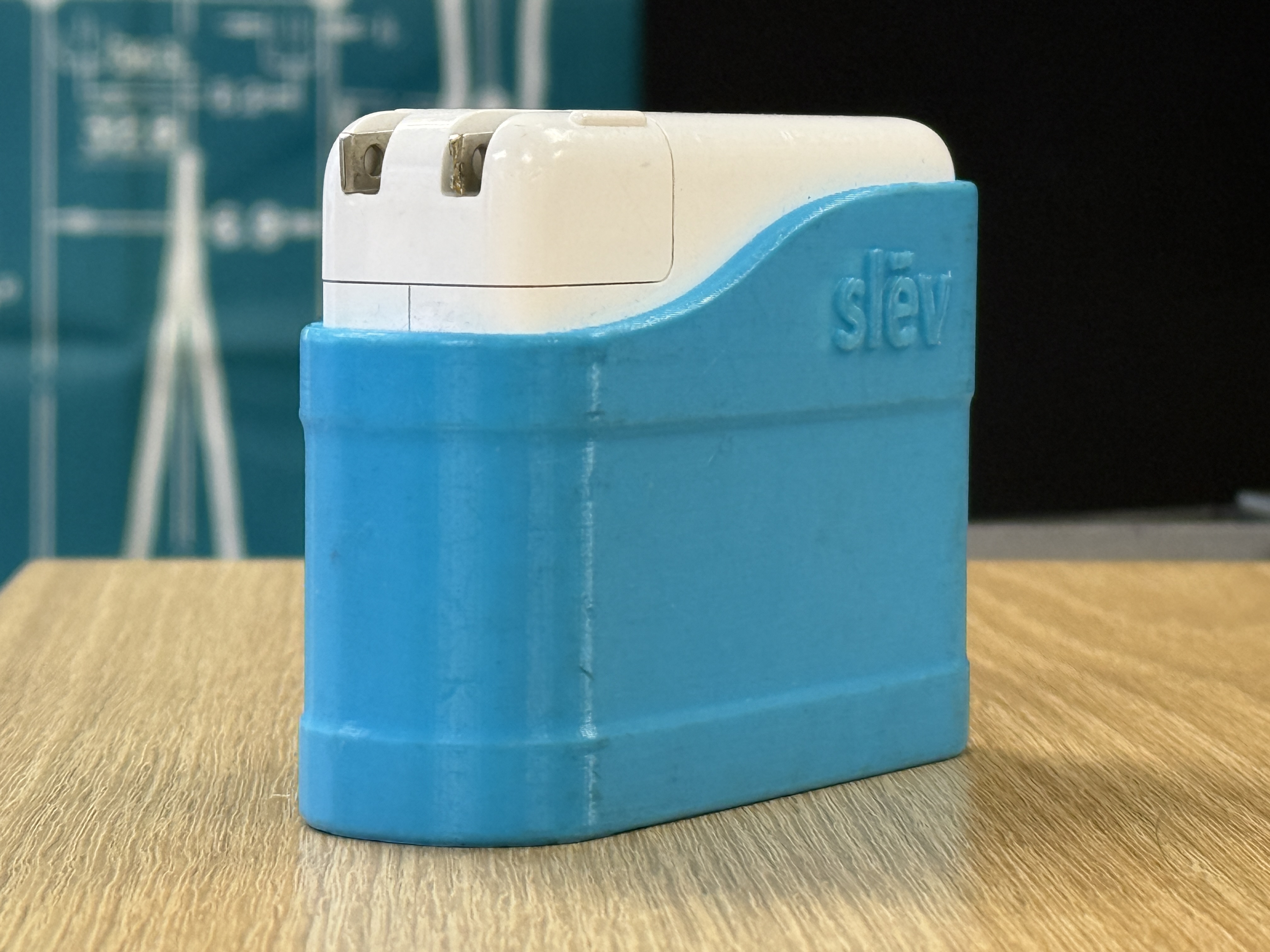 A small white Apple USB-C power adapter with its prongs visible at the top, wrapped from the lower two-thirds by a 3D-printed sleeve in sky blue PLA. The sleeve's profile rises in a curve to meet the white of the adapter. The word 'slēv' is embossed in lowercase into the sleeve's side, with a macron over the e. Horizontal FDM layer lines are visible across the blue surface. The adapter sits on a pale wood surface with out-of-focus technical drawings in the background.