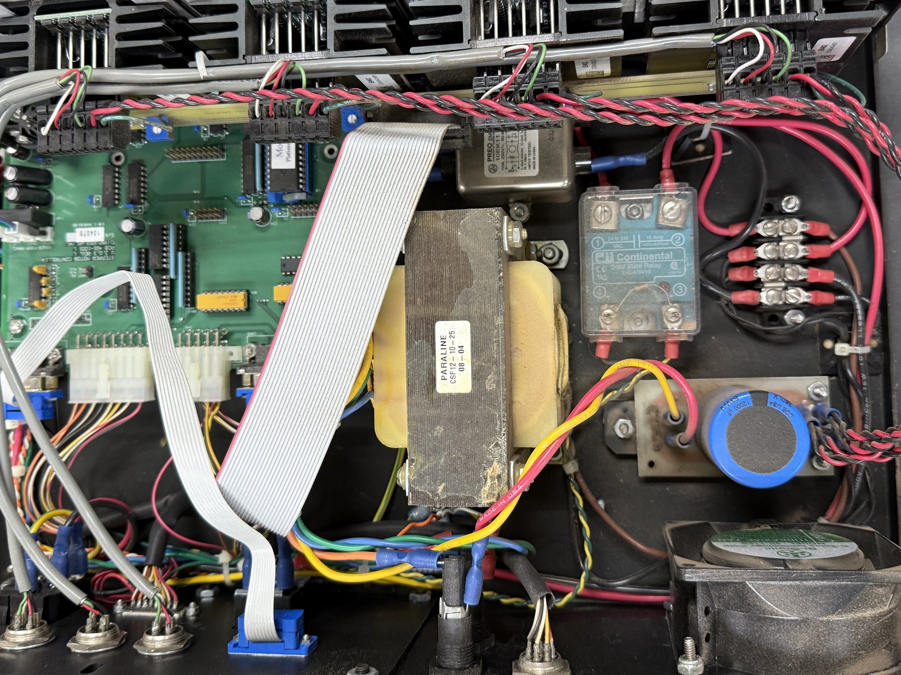 The inside of an electrical control cabinet with its cover removed. A green circuit board of DIP-package chips, trimmers and pin headers fills the left half; a grey ribbon cable arcs across it into a blue connector at the bottom edge. Center, a laminated-iron transformer with yellow bobbin windings, labeled PARALINE CSF12-10-25. Upper center, a metal-cased EMI line filter with its own schematic printed on the label. Right, a translucent CII Continental solid state relay bolted to the black mounting plate, a barrier terminal strip with stacked red ring terminals, a blue Cornell Dubilier capacitor, and an axial cooling fan. A tightly twisted red-and-black wire pair runs the full width of the frame along the top.