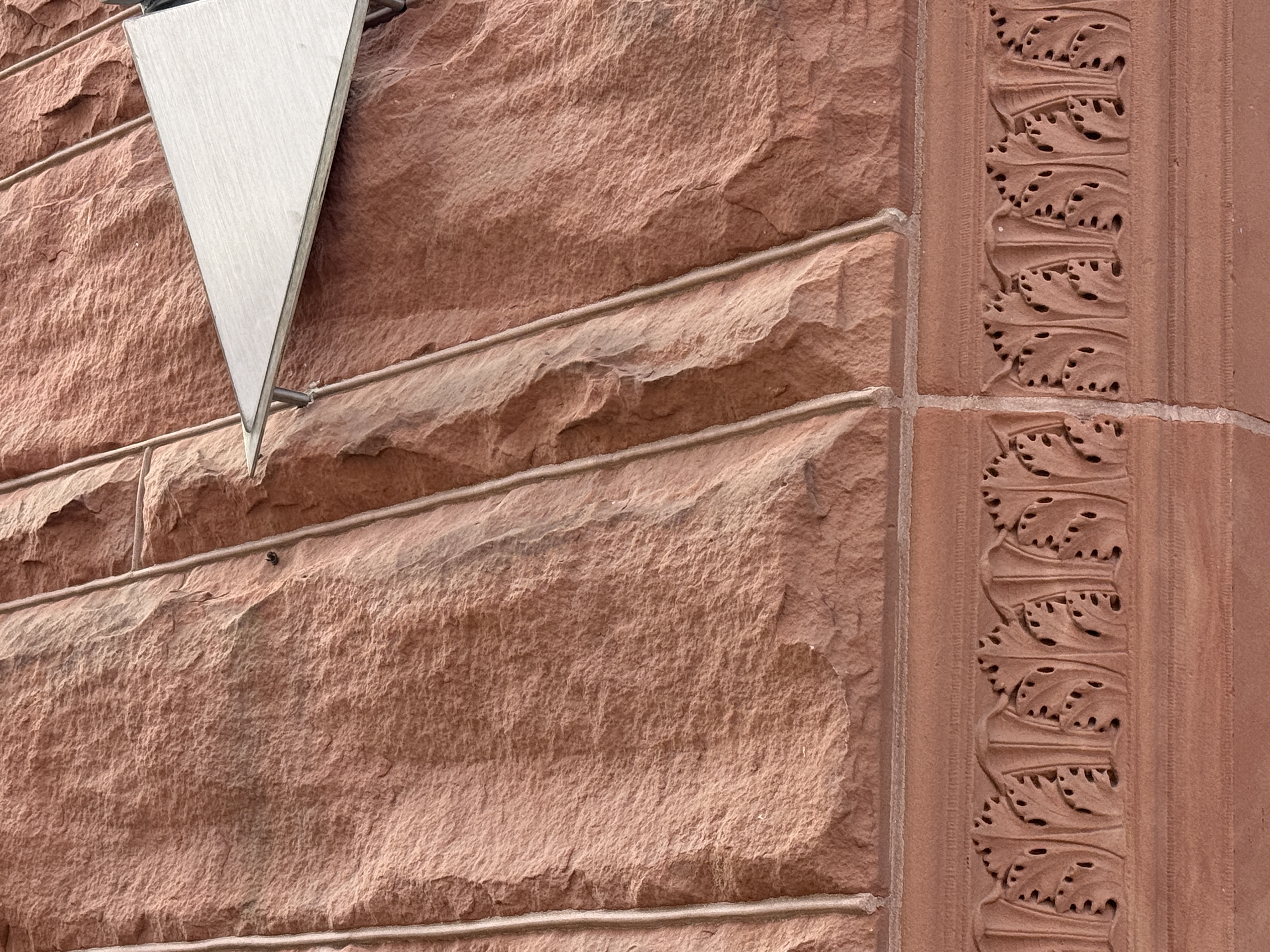 A close-up of a red sandstone building wall with rusticated rock-faced ashlar on the left and a carved vertical band of stylized leaves on the right.