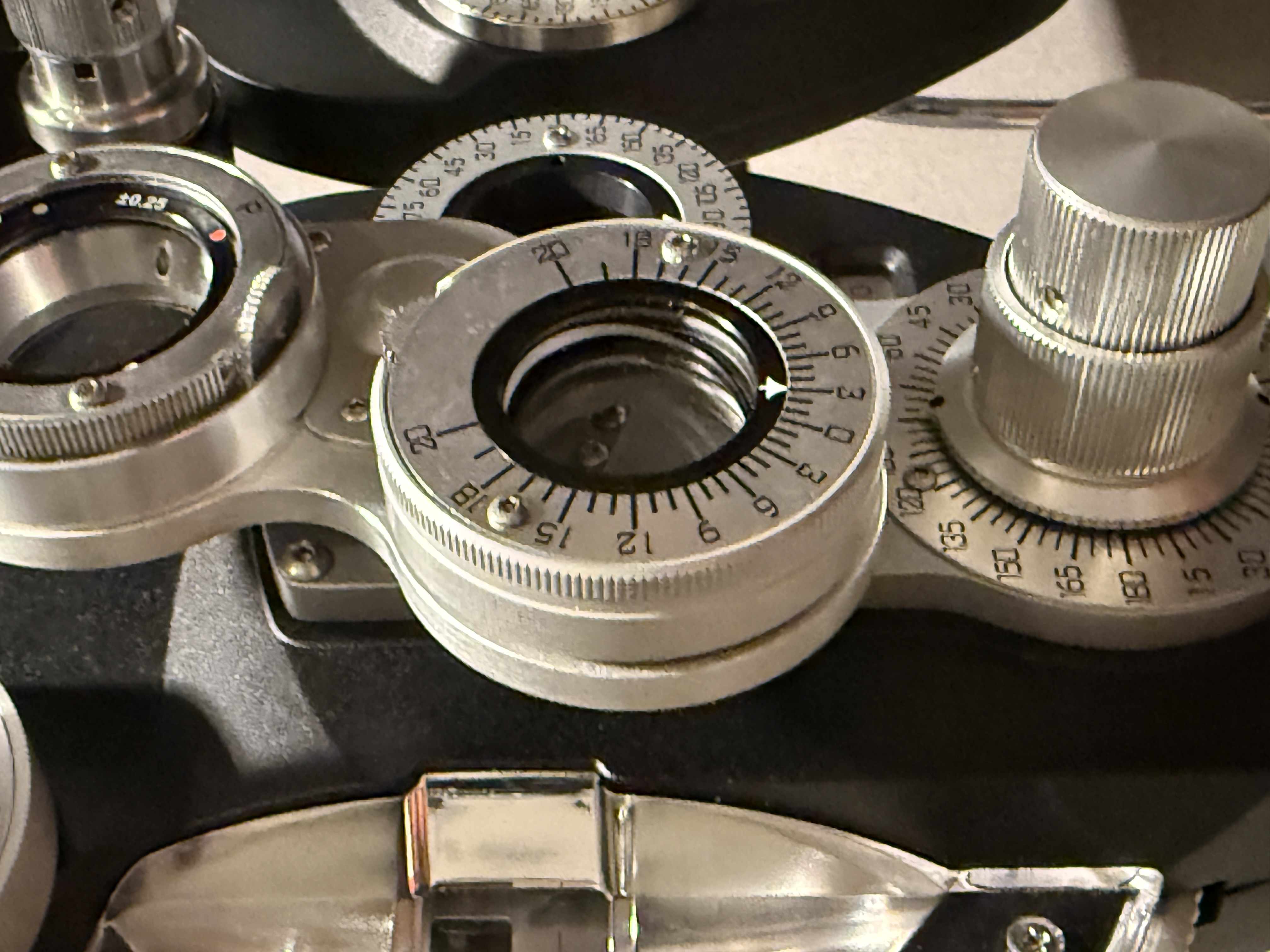 A close-up of a phoropter dial group, photographed from the operator's side. A Risley rotary prism sits at the center with a small viewing aperture; its circumference is engraved in symmetric pairs reading 20-18-15-12-9-6-3-0-3-6-9-12-15-18-20. Two arcs above and below carry axis-degree markings from 15 to 165 in five-degree graduations. Knurled chrome control knobs flank the assembly. A polished prism cube edges into the frame on the left.
