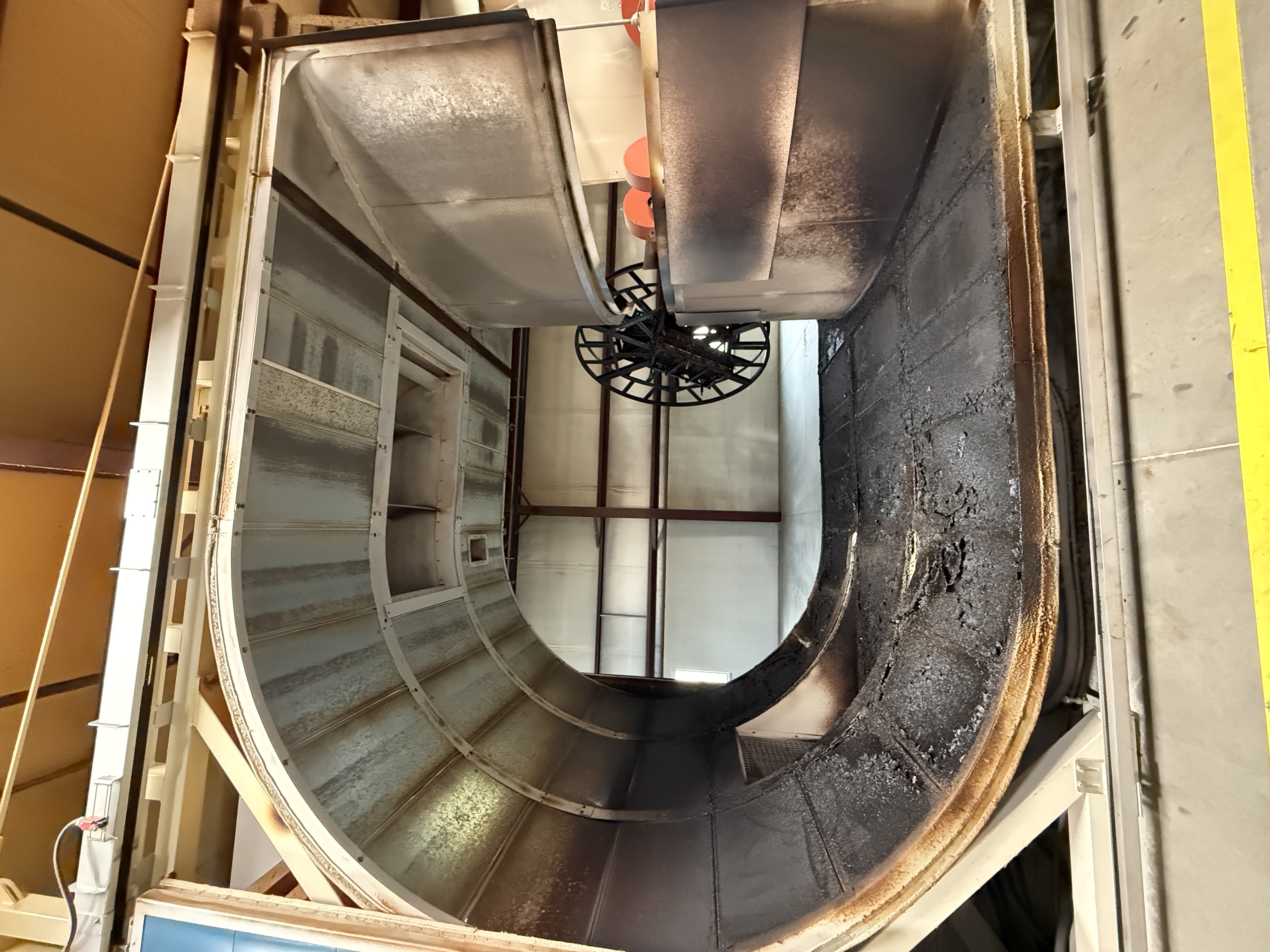 The interior of a biaxial rotational-molding oven seen through its open door — galvanized sheet above, carbonized black deposit below, a spoked spider frame visible in the factory beyond.