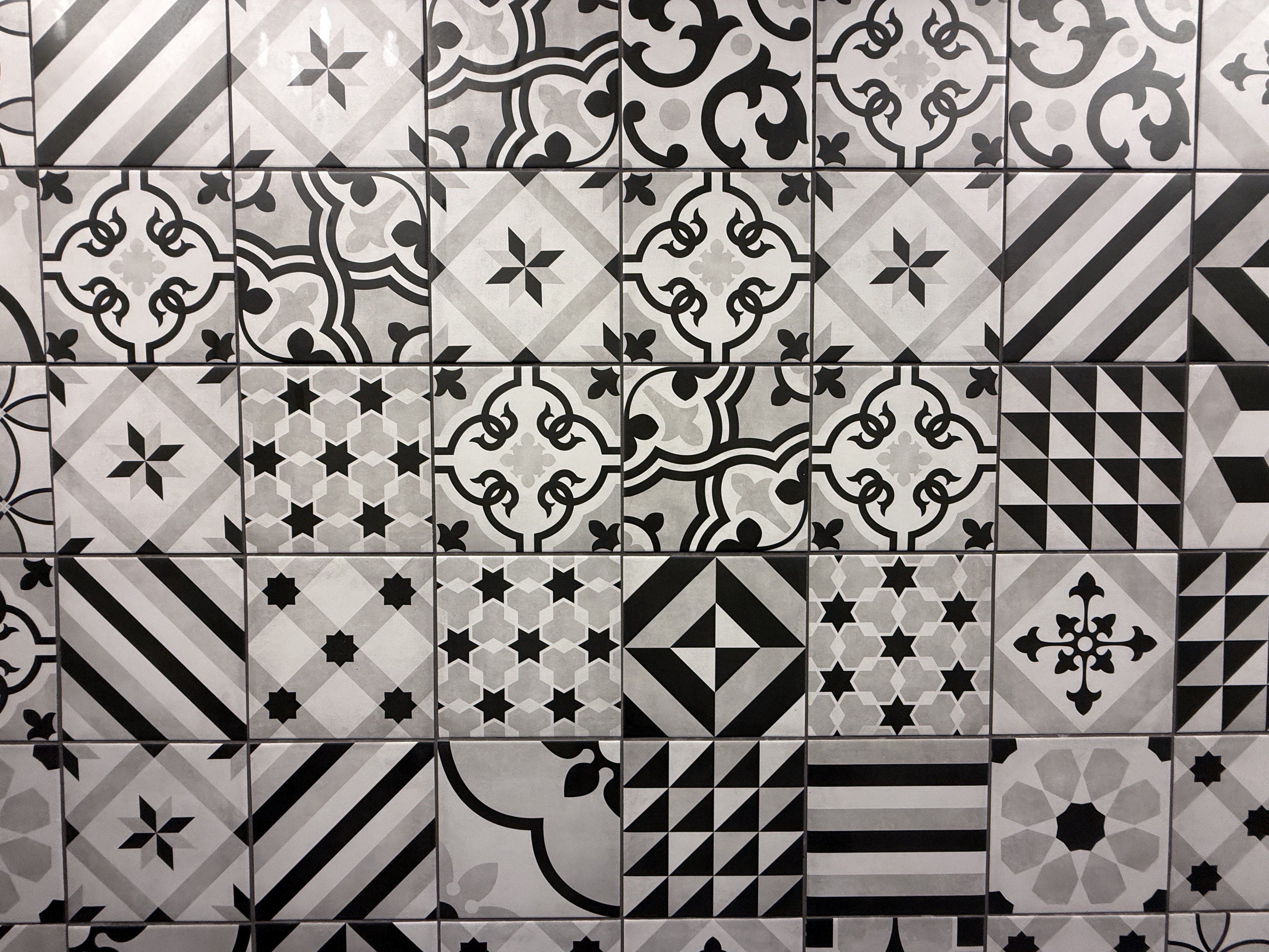 A wall of black-and-white patchwork tiles, square format, machine-cut. Roughly two dozen different patterns are visible &mdash; six-pointed stars in gray hex fields, black quatrefoils with ivy corners, diagonal stripes, foliate motifs, fields of small stars, lone geometric runs &mdash; laid in a perfectly regular grid with dead-straight grout. Several patterns recur across the wall.