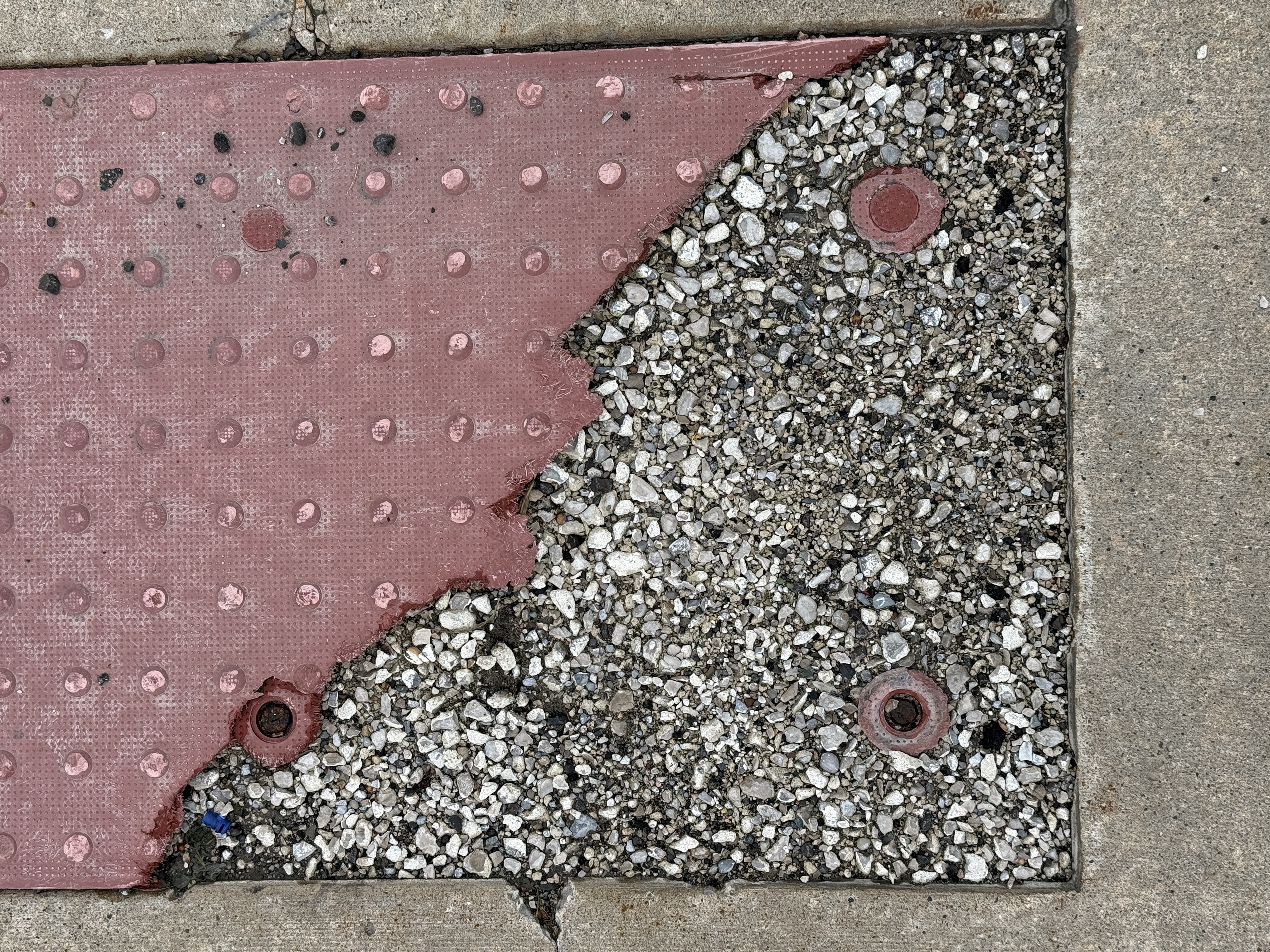 A damaged red tactile curb-ramp tile with its domed surface torn away, exposing embedded aggregate concrete and rusted fastener bosses.