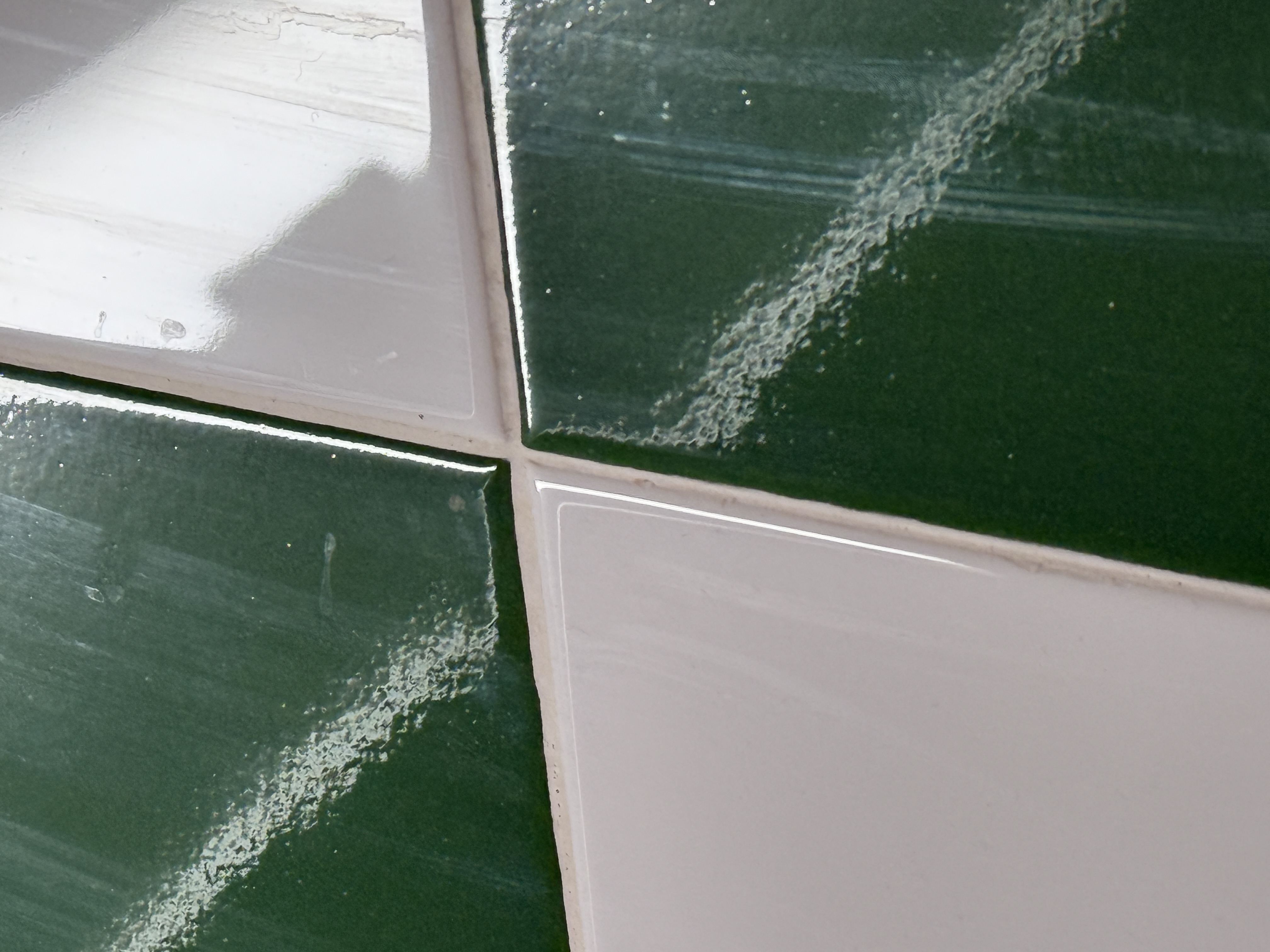 Four square glossy ceramic wall tiles meeting at a grout cross — two dark green, two white. The photograph is framed so the grid reads diagonally. A raking diagonal streak of lighter reflection cuts across both green tiles where the glaze has an orange-peel texture.