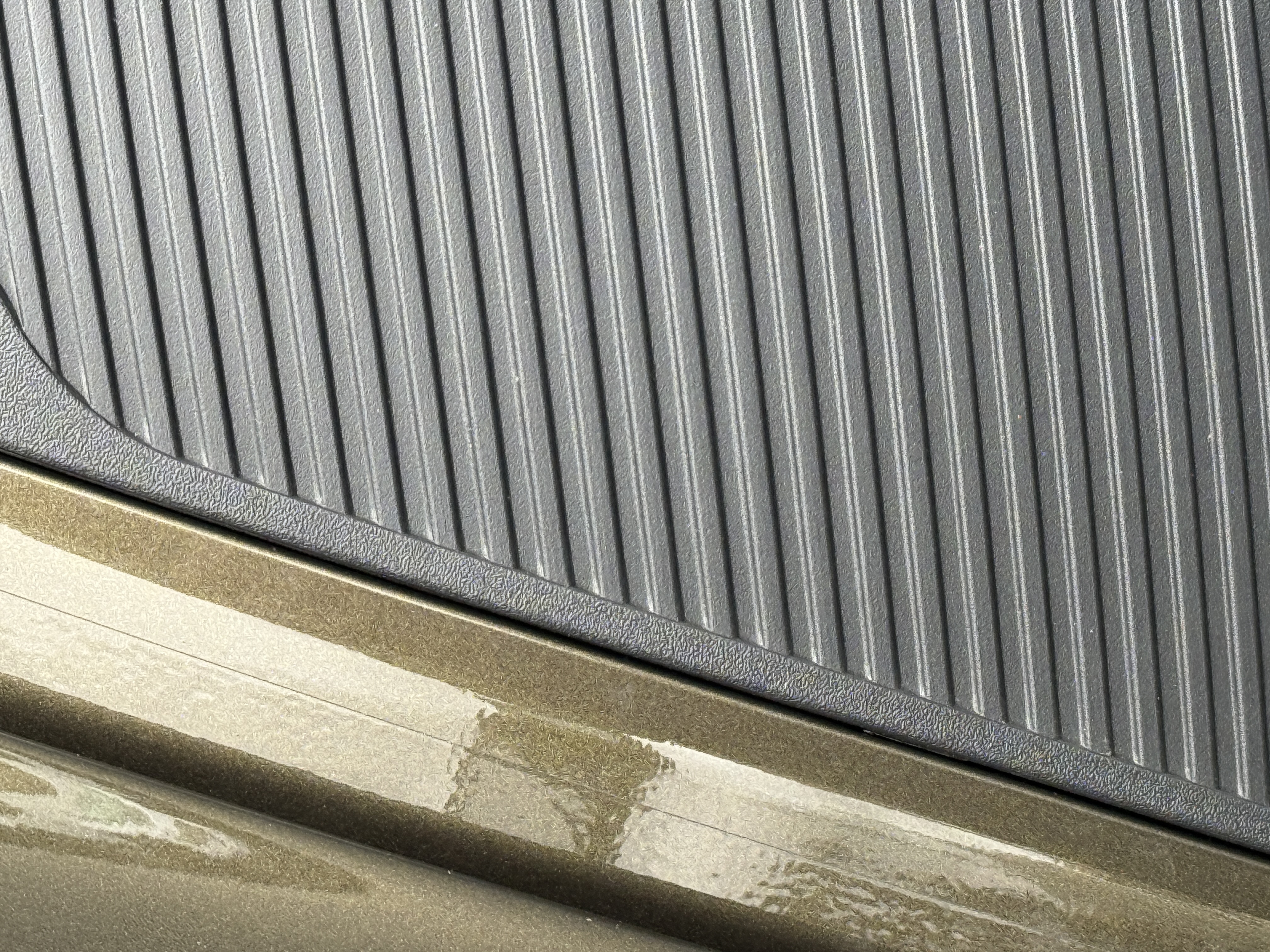 Two adjacent surfaces meeting at a vertical seam. On the left, a smooth olive-bronze painted or coated profile with what reads as a squeegee-mark of drying material. On the right, a dark panel with dense fine horizontal ribbing, curving slightly to follow an adjacent form.
